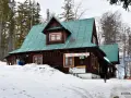Drewniany domek z zielonym dachem na przełęczy Przegibek, otoczony ośnieżonym lasem. Na ścianie wisi baner "BĘDZIE DOBRZE".