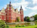 Zamek Mużakowski, okazała czerwona budowla z wieżami, otoczona zielenią i stawem, prezentuje się majestatycznie w słoneczny dzień.