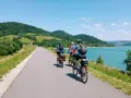 Trzy osoby na rowerach przemierzają malowniczą velo czorsztyn trasę wzdłuż jeziora, otoczone zielonymi wzgórzami.