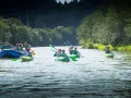 Grupa ludzi na tratwie i kajakach płynie rzeką wśród zielonych drzew.