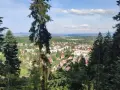 Widok na miasteczko zalesionej doliny, jakby z piekielnej góry. Słońce przebija się przez chmury, oświetlając dachy domów i zielone drzewa.