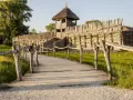 Drewniany mostek prowadzi do zrekonstruowanej osady Biskupin. Widać wał obronny, wieżę strażniczą i chaty kryte strzechą.