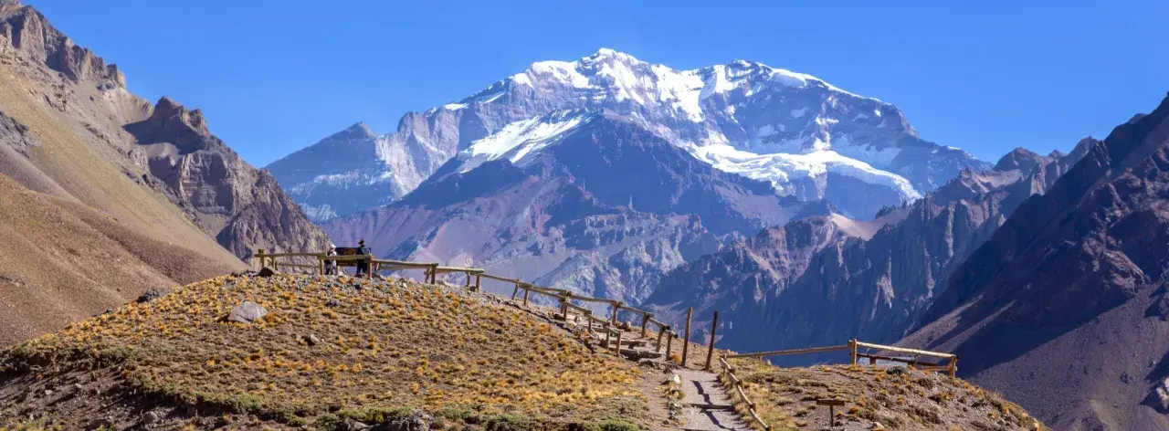 Widok na ośnieżony szczyt Aconcagua, cel wyprawy. Ścieżka z drewnianym ogrodzeniem prowadzi przez suchą, trawiastą górę.