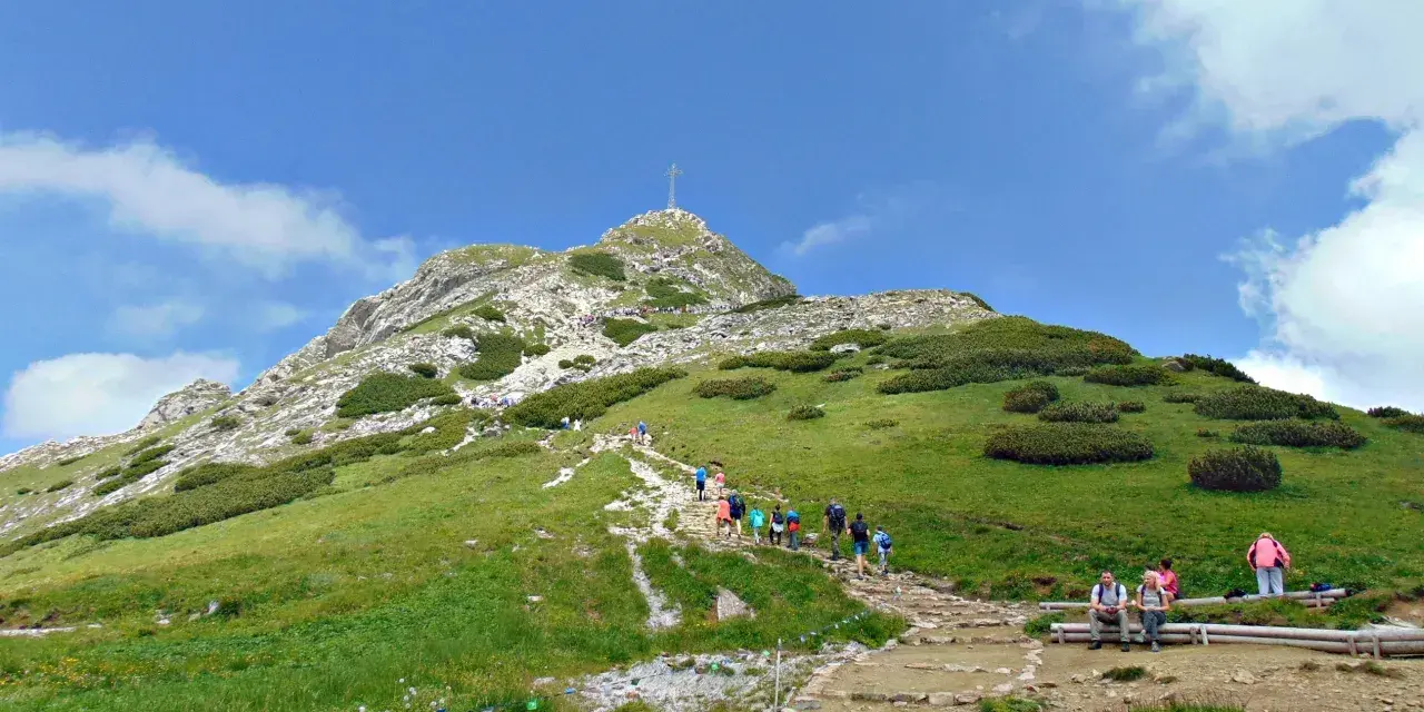 Tłum turystów wspina się kamienistym szlakiem na Giewont. Widać krzyż na szczycie.