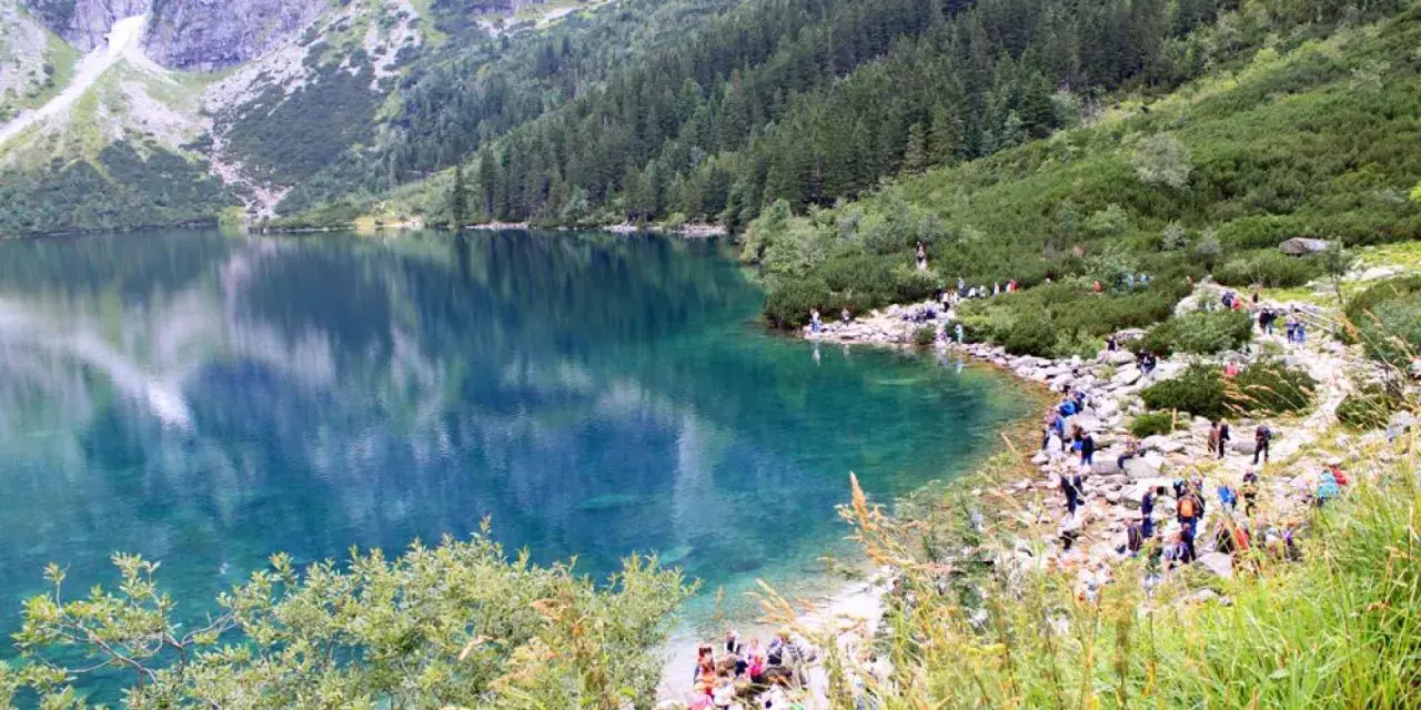 Turkusowe Morskie Oko otoczone zielonymi zboczami i skalistymi szczytami. Turyści spacerują brzegiem jeziora.