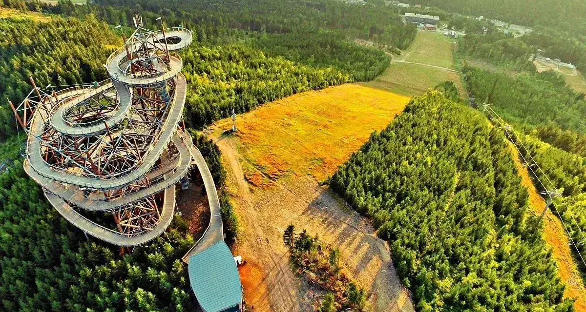 Widok z lotu ptaka na drewnianą wieżę widokową, najdłuższy most w Czechach, otoczoną lasem i złotym polem.