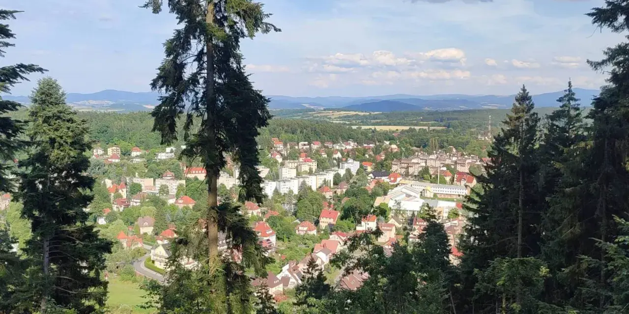 Widok na miasteczko zalesionej doliny, jakby z piekielnej góry. Słońce przebija się przez chmury, oświetlając dachy domów i zielone drzewa.
