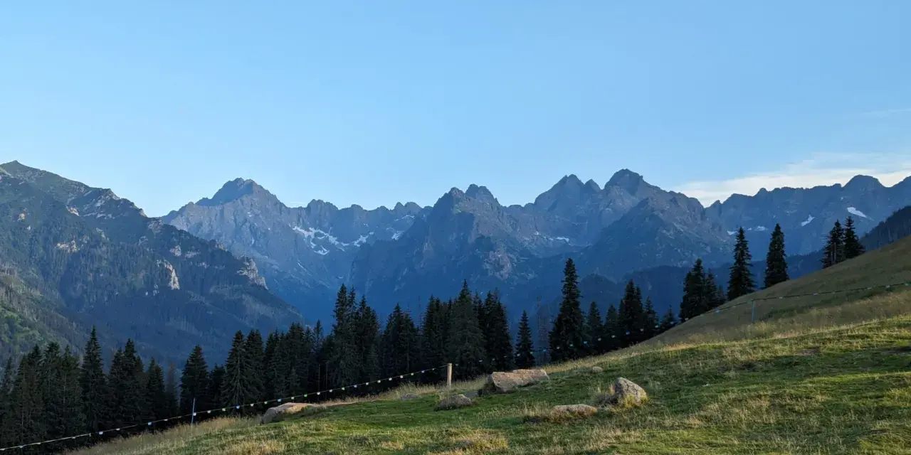 Widok na Tatry z zielonej łąki. Góry przypominają gęsią szyję, a ścieżka na zboczu sugeruje, ile schodów czeka na turystów.