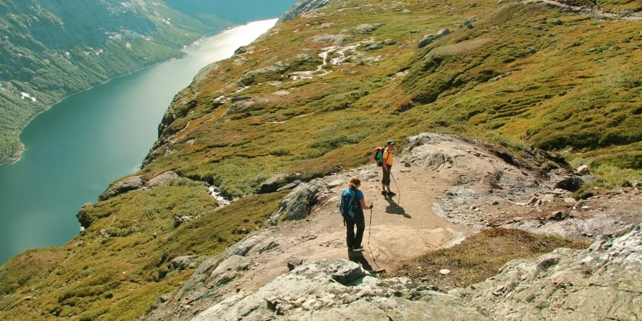 Dwie osoby wędrują po skalistym szlaku w górach Norwegii, podziwiając malowniczy fiord.