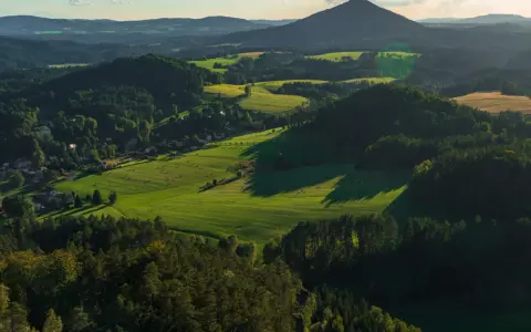 Malowniczy krajobraz Czech z zielonymi polami, lasami i górami w oddali. Słońce rzuca długie cienie na zbocza.