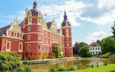 Zamek Mużakowski, okazała czerwona budowla z wieżami, otoczona zielenią i stawem, prezentuje się majestatycznie w słoneczny dzień.