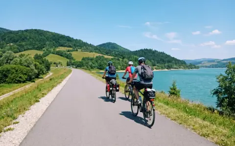 Trzy osoby na rowerach przemierzają malowniczą velo czorsztyn trasę wzdłuż jeziora, otoczone zielonymi wzgórzami.