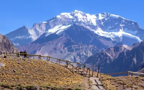 Widok na ośnieżony szczyt Aconcagua, cel wyprawy. Ścieżka z drewnianym ogrodzeniem prowadzi przez suchą, trawiastą górę.