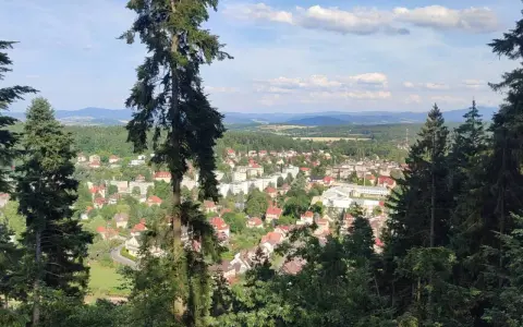 Widok na miasteczko zalesionej doliny, jakby z piekielnej góry. Słońce przebija się przez chmury, oświetlając dachy domów i zielone drzewa.