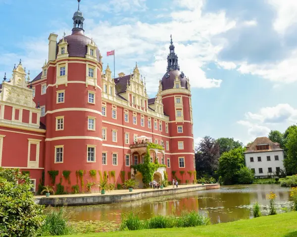 Zamek Mużakowski, okazała czerwona budowla z wieżami, otoczona zielenią i stawem, prezentuje się majestatycznie w słoneczny dzień.