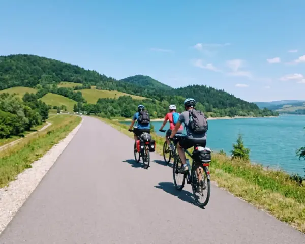 Trzy osoby na rowerach przemierzają malowniczą velo czorsztyn trasę wzdłuż jeziora, otoczone zielonymi wzgórzami.