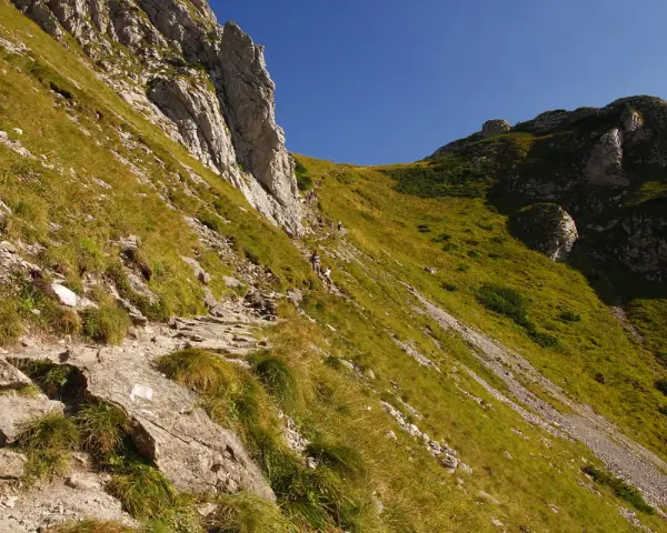 Kamienista ścieżka, czerwony szlak na Giewont, wije się po zboczu porośniętym trawą. W oddali widać turystów.