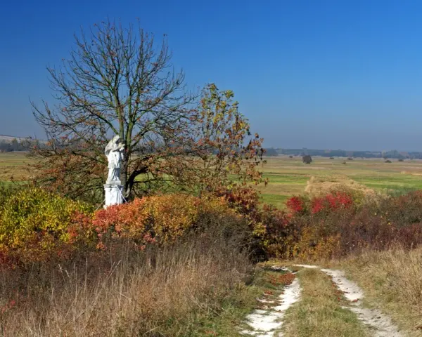 Kamienna figura Matki Boskiej na tle jesiennego krajobrazu Ponidzia. Droga wśród traw prowadzi do kapliczki.