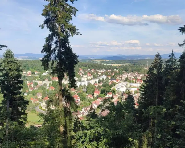 Widok na miasteczko zalesionej doliny, jakby z piekielnej góry. Słońce przebija się przez chmury, oświetlając dachy domów i zielone drzewa.
