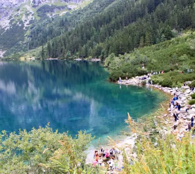 Turkusowe Morskie Oko otoczone zielonymi zboczami i skalistymi szczytami. Turyści spacerują brzegiem jeziora.