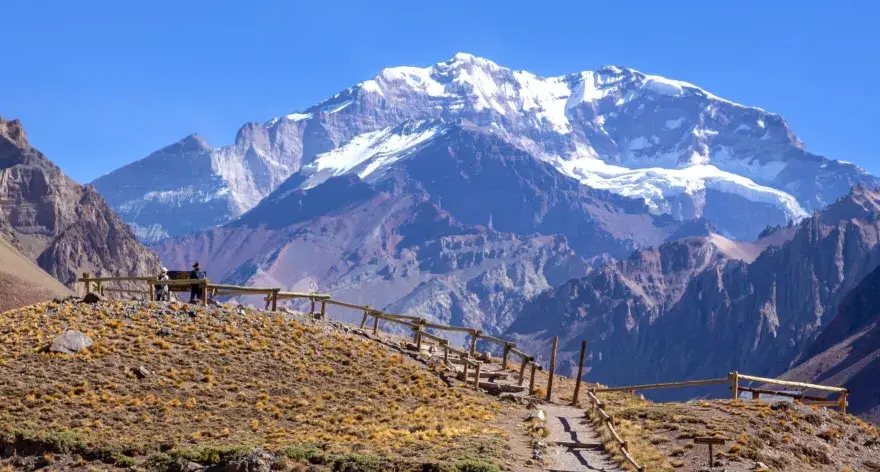 Widok na ośnieżony szczyt Aconcagua, cel wyprawy. Ścieżka z drewnianym ogrodzeniem prowadzi przez suchą, trawiastą górę.