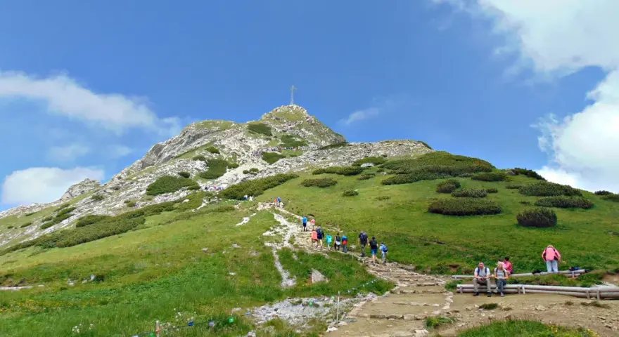 Tłum turystów wspina się kamienistym szlakiem na Giewont. Widać krzyż na szczycie.
