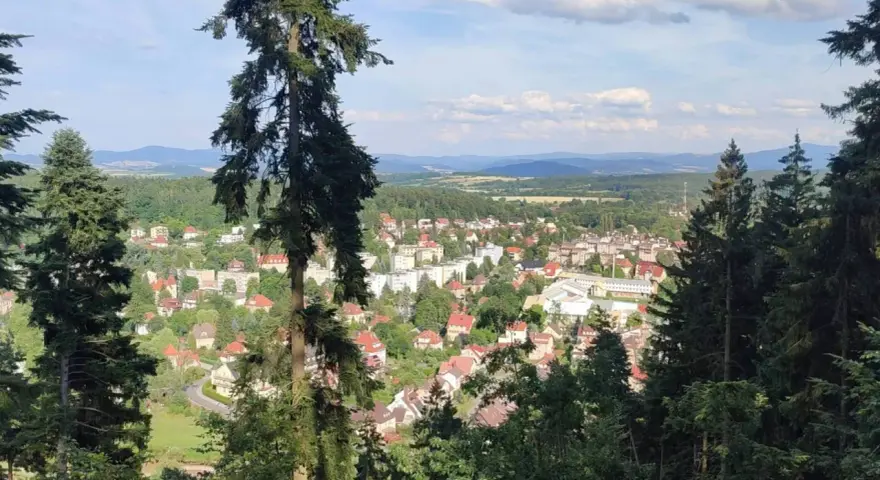 Widok na miasteczko zalesionej doliny, jakby z piekielnej góry. Słońce przebija się przez chmury, oświetlając dachy domów i zielone drzewa.