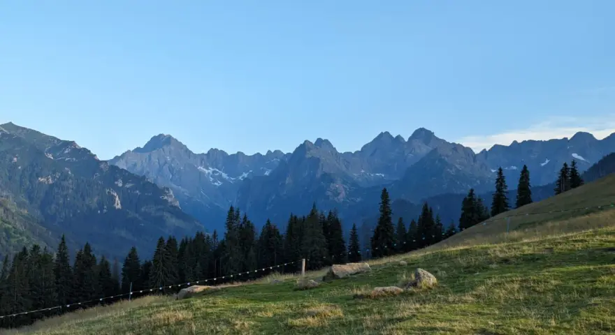 Widok na Tatry z zielonej łąki. Góry przypominają gęsią szyję, a ścieżka na zboczu sugeruje, ile schodów czeka na turystów.