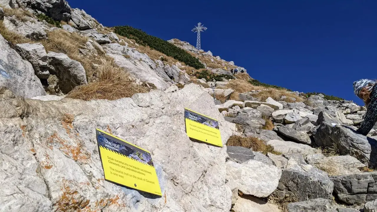 Kamienista ścieżka na Giewont. Tabliczki ostrzegają, że szlak jest jednokierunkowy. W oddali widać krzyż na szczycie.
