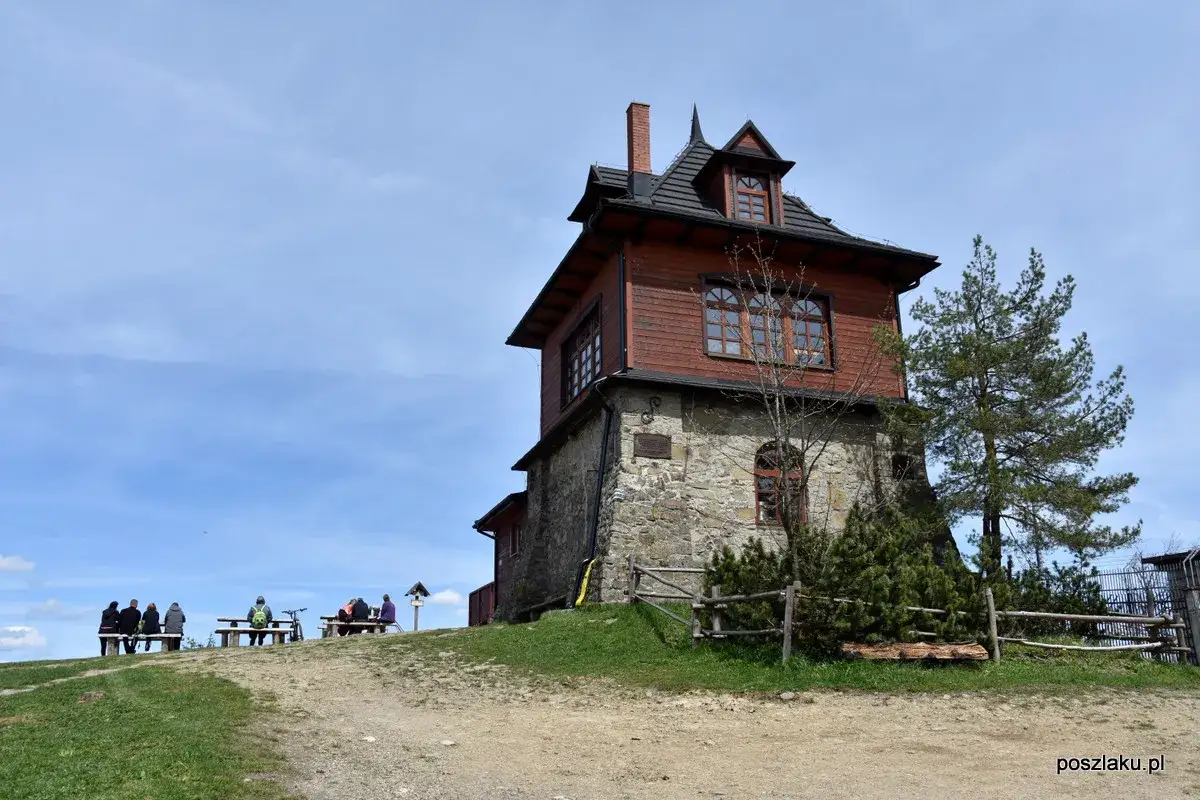 Drewniane schronisko na szlaku Luboń Wielki. Ludzie odpoczywają na ławkach, podziwiając widok.