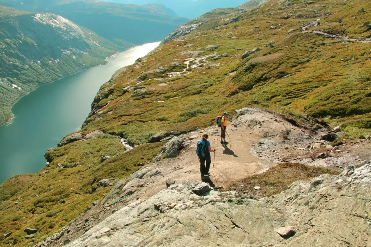 Dwie osoby wędrują po skalistym szlaku w górach Norwegii, podziwiając malowniczy fiord.