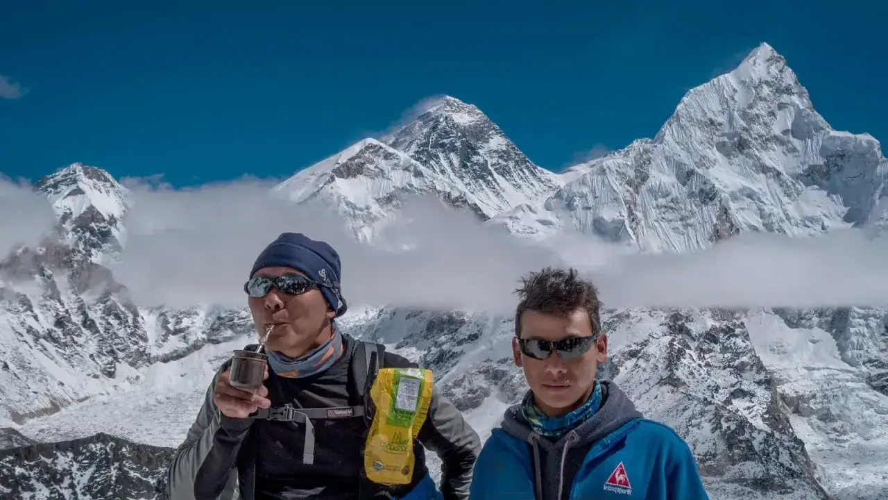 Dwóch alpinistów na tle majestatycznego szczytu Mount Everest. Jeden pije yerba mate, drugi patrzy w obiektyw. Widok zapiera dech w piersiach.