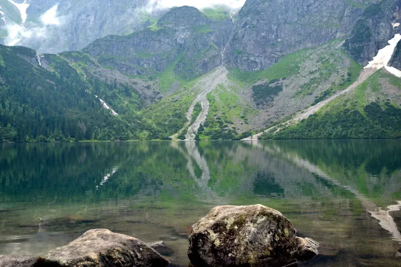 Spokojne Morskie Oko, odbijające skaliste zbocza i zielone lasy. Palenica Białczańska w oddali.