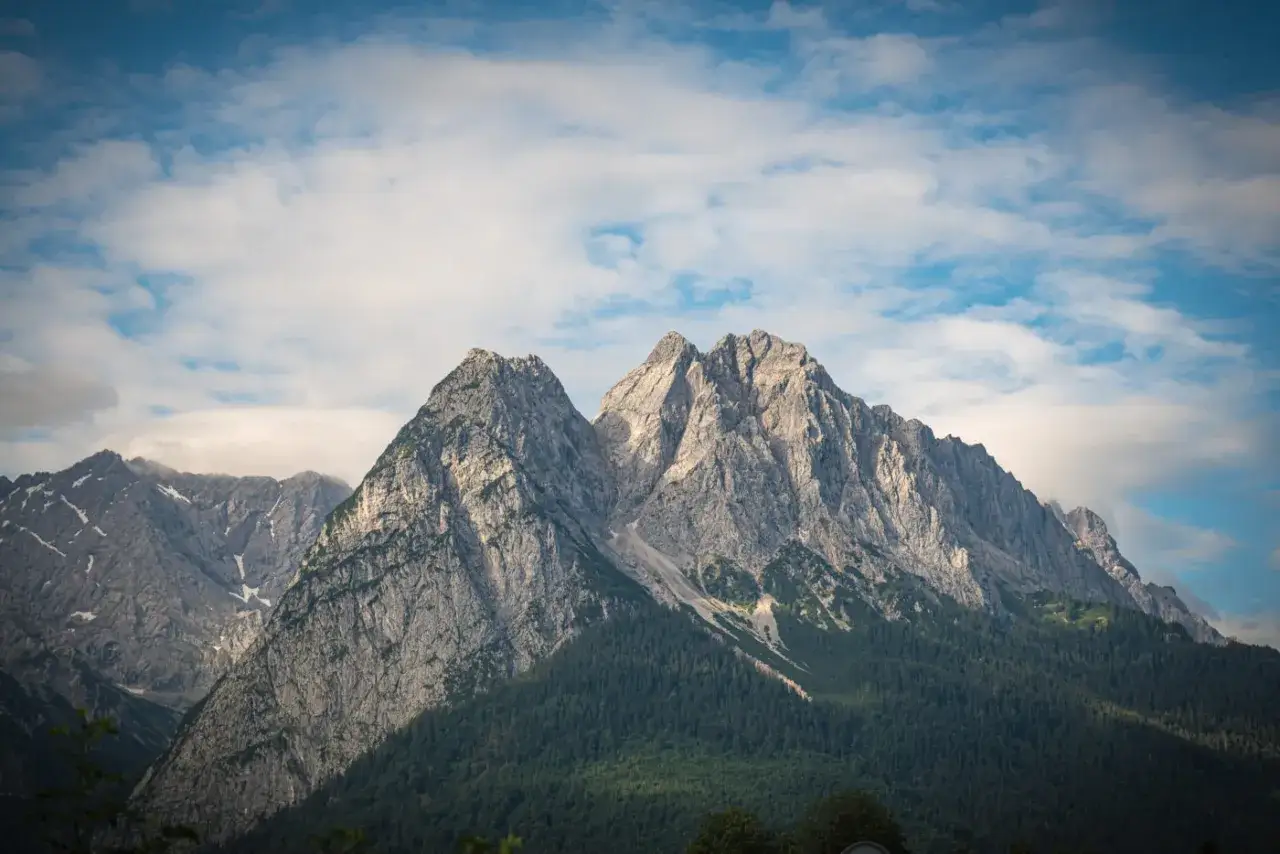 Majestatyczne szczyty górskie, w tym najwyższy szczyt Niemiec, Zugspitze, wznoszą się na tle błękitnego nieba z chmurami.