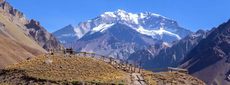 Widok na ośnieżony szczyt Aconcagua, cel wyprawy. Ścieżka z drewnianym ogrodzeniem prowadzi przez suchą, trawiastą górę.