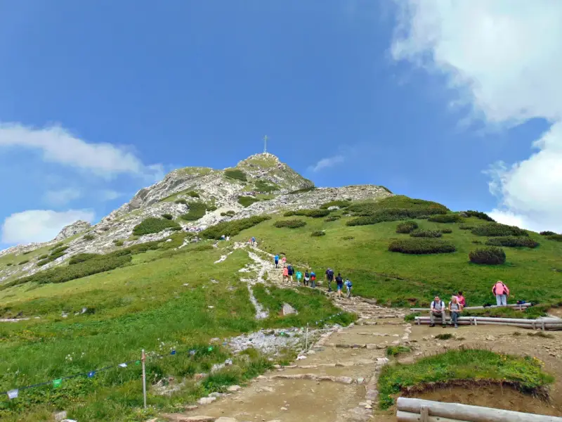 Tłum turystów wspina się kamienistym szlakiem na Giewont. Widać krzyż na szczycie.